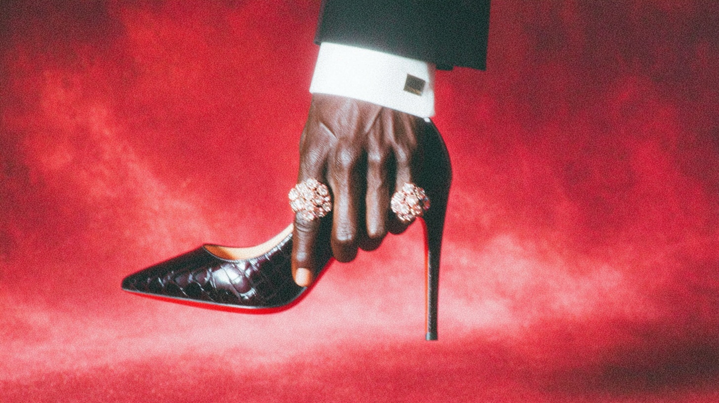 AI-generated editorial image by Maria Lankina depicting a hand holding a red high heel against a crimson backdrop - symbolic study of power, authorship, and hierarchy accompanying the Mythohuman Lens essay “Red as Power, Red as Skin.”